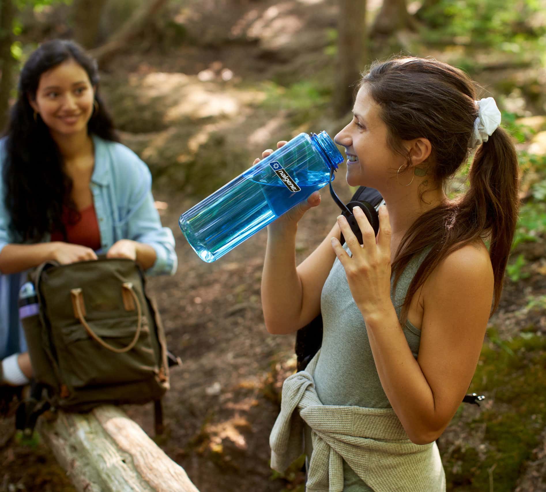 Young woman sips from a blue Nalgene 48oz water bottle while on a hike with a friend